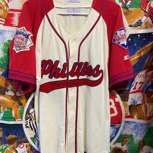 May include: Cream-colored baseball jersey with red sleeves and trim. Features the word "Phillies" in red script across the chest, with a "National League" patch on the left sleeve and a "Phillies" patch on the right sleeve.