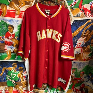 Könnte beinhalten: Ein rotes Hawks-Baseballtrikot mit weiß-gelben Verzierungen. Das Wort "Hawks" steht in einer verblassten Schriftart auf der Brust. Das Trikot hat eine Knopfleiste und ein Hawks-Logo auf dem Ärmel.