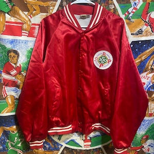 Pode incluir: Casaco bomber vermelho acetinado com acabamentos listrados brancos e vermelhos nos punhos e na cintura. O casaco tem uma gola branca e um patch circular no peito com o texto "100" e "1983".