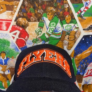Op de afbeelding: Een marineblauwe baseballpet met het woord "FLYERS" in oranje geborduurd. De hoed heeft een oranje rand en een gebogen klep. De achtergrond heeft een sportthema.