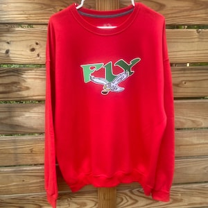 May include: A red long-sleeve sweatshirt with a green and white graphic of an eagle and the word "FLY". The sweatshirt is hanging on a white hanger against a wooden background.
