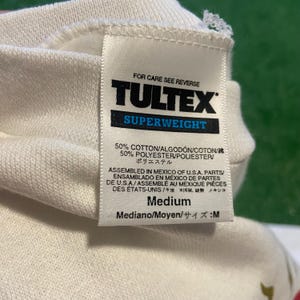 May include: Close-up of a white TULTEX Superweight tag. The tag includes text in English, Spanish, and French, with the size listed as Medium. The tag indicates the shirt is made of 50% cotton and 50% polyester.