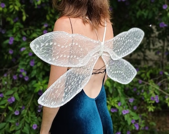 May include: A person wearing a teal blue velvet dress with white lace butterfly wings. The person is standing in front of a green bush with purple flowers.