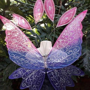 Puede incluir: Un par de alas de hada iridiscentes con un diseño degradado de rosa y morado. Las alas están decoradas con purpurina plateada y remolinos.