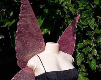 May include: Pink fairy wings with a swirl design and glitter. The wings are made of sheer fabric and are attached to a white strap.