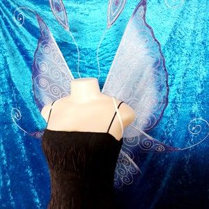 Peut inclure: Une robe noire à fines bretelles et une paire d'ailes de fée. Les ailes sont bleu clair avec une bordure bleu foncé et des volutes argentées. Les ailes sont fixées à un bandeau blanc avec deux petites antennes décoratives.