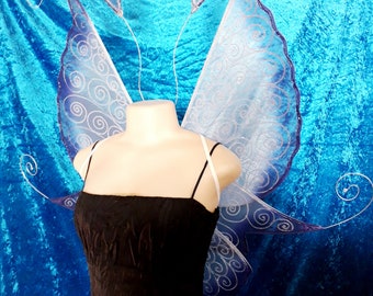 Könnte beinhalten: Ein schwarzes Kleid mit dünnen Trägern und ein Paar Feenflügel. Die Flügel sind hellblau mit dunkelblauem Rand und silbernen Wirbeln. Die Flügel sind an einem weißen Stirnband mit zwei kleinen, dekorativen Antennen befestigt.