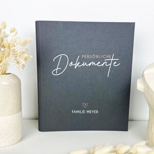 Könnte beinhalten: Ein schwarzer Ordner mit dem Text "Persönliche Dokumente" in weißer Kursivschrift und "Familie Meyer" in weißer Schrift mit einem rosa Herz darüber.