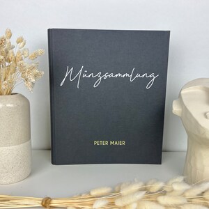 Könnte beinhalten: Ein schwarzes Hardcover-Journal mit dem Text "Münzsammlung" in weißer Kursivschrift und "PETER MAIER" in goldenen Lettern. Das Journal liegt auf einem Tisch mit einer Vase mit Trockenblumen und einer weißen Keramikskulptur.