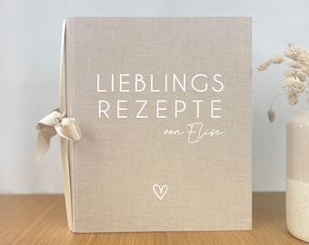 May include: A beige linen-covered recipe book with the words "LIEBLINGS REZEPTE von Elise" in white. A small white heart is below the text. The book is tied with a cream-colored ribbon. A vase with dried flowers is in the background.