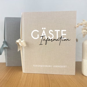 Könnte beinhalten: Ein beigefarbener Gästeinformationsordner aus Leinen mit einem weißen Band darum gebunden. Der Ordner trägt den Text "GÄSTE Information" in weißen Buchstaben und "FERIENWOHNUNG LEBENSWERT" in schwarzen Buchstaben.