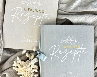 May include: Two gray recipe books with a white floral design and the words "Lieblings Rezepte" and "Familien Rezepte" in gold and white lettering. The books are tied together with a blue ribbon and the names "Sabrina" and "Daniel" are written on the front of each book.