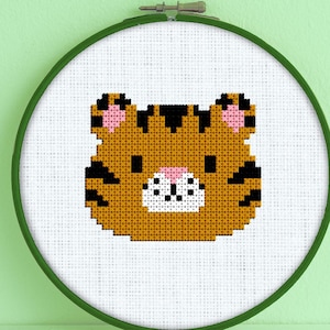 May include: A cross-stitch embroidery hoop with a cute tiger face design. The tiger is orange with black stripes and pink ears. The hoop is green.