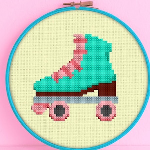 Op de afbeelding: Een kruissteekpatroon van een turquoise rolsschaats met roze veters en wielen op een crèmekleurige achtergrond. Het patroon is ingelijst in een blauwe hoepel.