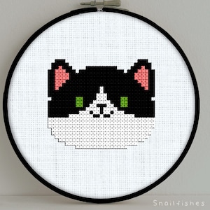 Peut inclure: Modèle de point de croix de chat pixelisé noir et blanc avec des oreilles roses et des yeux verts. Le motif est dans un cercle de broderie rond.
