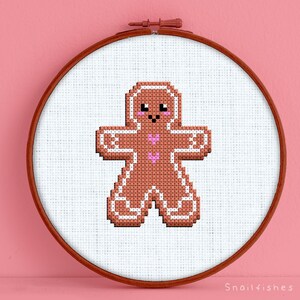 May include: A brown gingerbread man cross stitch pattern with pink hearts on the chest, framed in a wooden embroidery hoop. The hoop is on a pink background. "Snailfishes Stitches" is written in white text at the bottom of the image.