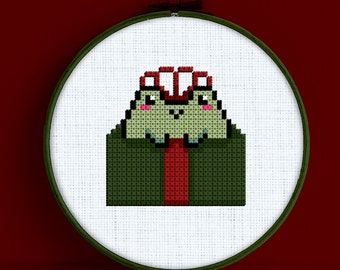 May include: A green and red cross-stitch embroidery of a frog wearing a red hat and a red tie. The embroidery is framed in a white hoop with a green outer ring. The text "Snailfishes Stitches" is printed below the hoop.