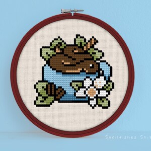 Könnte beinhalten: Ein Kreuzstich-Stickrahmen mit einem braunen und blauen Design eines Bären in einer blauen Tasse mit einer weißen Blume und grünen Blättern. Der Text "Snailfishes Stitches" befindet sich am unteren Rand des Rahmens.