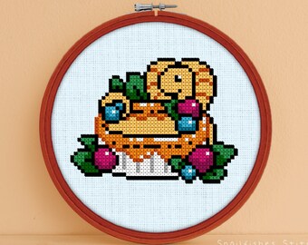 Peut inclure: Motif de point de croix représentant un chat jaune et orange assis dans un bol de fruits. Le bol est rempli de myrtilles, de framboises et de feuilles vertes. Le chat regarde le spectateur. Le motif est encadré dans un cercle en bois.