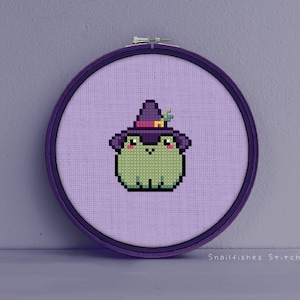 Puede incluir: Un patrón de punto de cruz de una rana verde que lleva un sombrero de bruja morado con una flor amarilla en la parte superior. La rana está sobre un fondo morado claro.