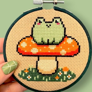 May include: Cross-stitch embroidery in a wooden hoop. The design features a green frog sitting on a red and orange mushroom with white spots. Green grass and small flowers are at the base of the mushroom. The hoop is held by a hand.