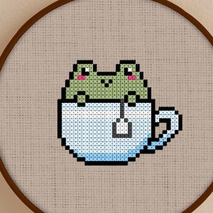 Puede incluir: Un aro de bordado de punto de cruz con una rana verde sentada en una taza de té azul con una bolsita de té. La rana tiene mejillas rosadas y mira al espectador. El aro está sobre una superficie blanca.