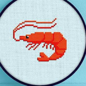 May include: A white embroidery hoop with a red shrimp cross-stitch design. The shrimp is curled with its tail up and its head facing left. The hoop is on a light blue background.