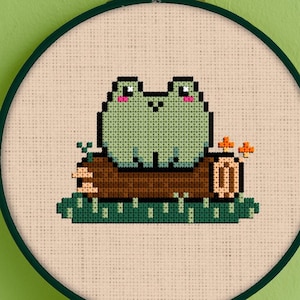 May include: A green frog with a pink nose and eyes sits on a brown log with green leaves and orange flowers. The frog is embroidered on a beige fabric with a brown wooden frame.