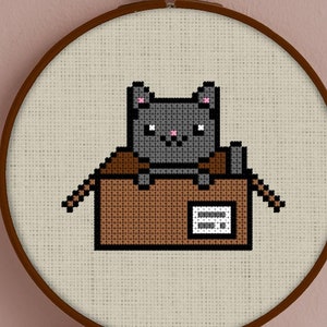 May include: A gray cat sits inside a brown cardboard box with a label. The cat is looking at the viewer. The box is on a white background.