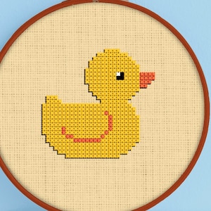 Puede incluir: Un patrón de punto de cruz de un pato de goma amarillo en un aro de bordado marrón. El pato está cosido con hilo amarillo, naranja y rojo.