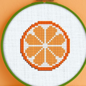 May include: A cross-stitch embroidery hoop with a white background and an orange citrus fruit design. The design is a simple, pixelated representation of an orange slice.