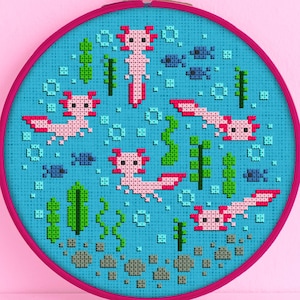 Könnte beinhalten: Ein blauer und rosa Kreuzstich-Stickrahmen mit einem Design von Axolotln, die in einem Teich mit grünen Pflanzen und Blasen schwimmen. Der Rahmen befindet sich auf einem rosa Hintergrund.