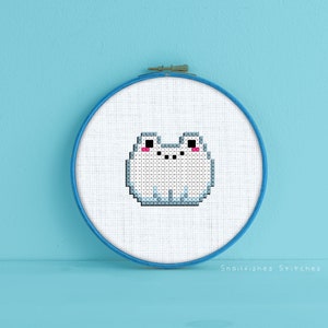 Op de afbeelding: Een witte kruissteek-borduurwerk van een cartoon kikker met roze wangen in een blauwe borduurring. De ring staat op een lichtblauwe ondergrond. "Snailfishes Stitches" staat in zwarte tekst onder de ring.