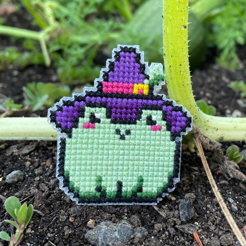 May include: A green frog wearing a purple witch hat with a pink and yellow band. The frog is embroidered with black eyes and a pink mouth. The frog is sitting on a bed of brown dirt.