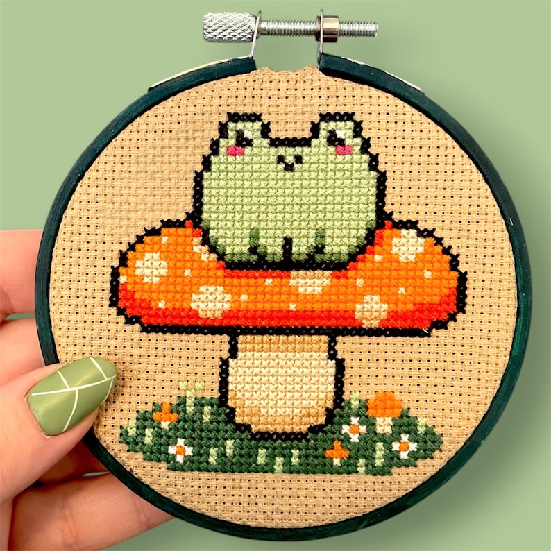 May include: Cross-stitch embroidery in a wooden hoop. The design features a green frog sitting on a red and orange mushroom with white spots. Green grass and small flowers are at the base of the mushroom. The hoop is held by a hand.