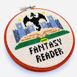 May include: A white embroidery hoop with a red wooden frame. The embroidery depicts a black dragon flying over a green book with blue castles and yellow stars. The text "Fantasy Reader" is stitched in black below the book.