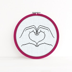 May include: A pink embroidery hoop with white fabric inside. The fabric has a black outline of two hands forming a heart shape.