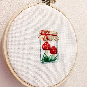 May include: A white embroidery hoop with a cross-stitch design of a glass jar filled with red and white mushrooms and green grass. The jar has a brown lid with a red ribbon.