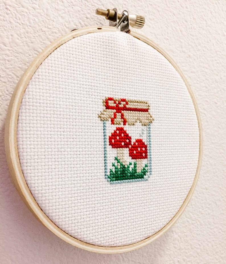 May include: A white embroidery hoop with a cross-stitch design of a glass jar filled with red and white mushrooms and green grass. The jar has a brown lid with a red ribbon.