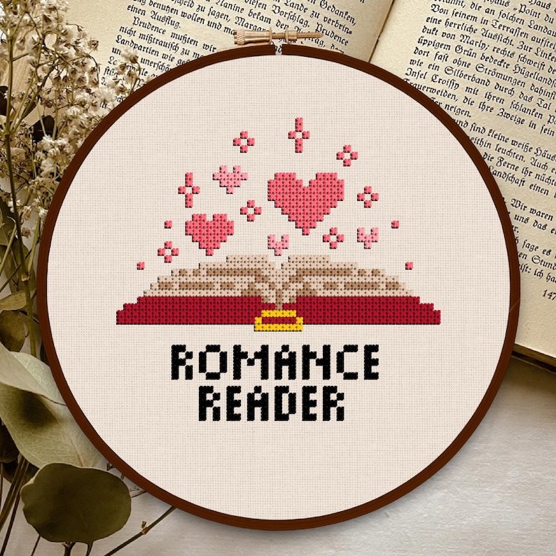 May include: A white embroidery hoop with a red and brown open book with a gold clasp. Pink hearts are floating above the book. The text "ROMANCE READER" is below the book.