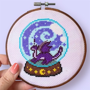 May include: Cross-stitch embroidery in a wooden hoop. The design features a purple dragon inside a snow globe with a swirling blue and purple background. The base of the snow globe is brown with yellow star and moon accents.