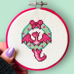 May include: A cross-stitch embroidery hoop with a pink flamingo design. The flamingo is encircled by a wreath of green leaves and pink flowers, with a pink bow at the top. The hoop is a bright pink colour.