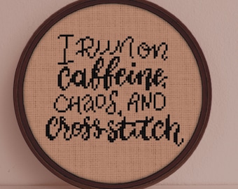 May include: A brown wooden embroidery hoop with beige fabric stretched across it. The fabric has black cross-stitch embroidery that reads "I run on caffeine, chaos, and cross-stitch."