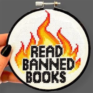 May include: A circular embroidery hoop containing a cross-stitch design on white fabric. The design features a stylized flame in shades of yellow, orange, and red, with the black text "READ BANNED BOOKS" centered prominently over the flames.
