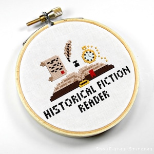 May include: Cross-stitch embroidery in a wooden hoop. The design features a book, scroll, quill, and pocket watch, with the words "HISTORICAL FICTION READER" in black. The embroidery is on white fabric.