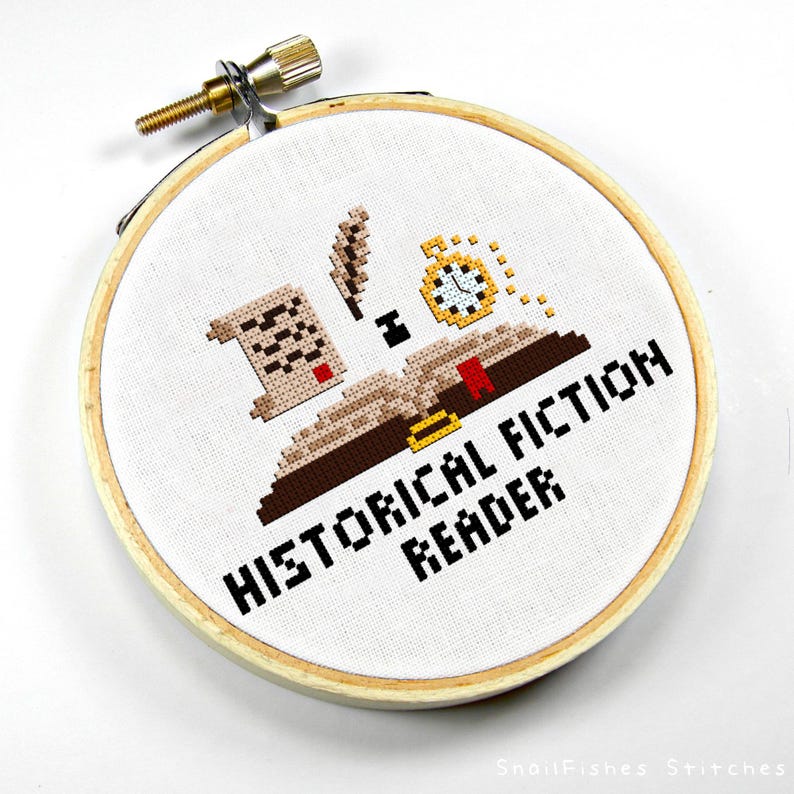 May include: Cross-stitch embroidery in a wooden hoop. The design features a book, scroll, quill, and pocket watch, with the words "HISTORICAL FICTION READER" in black. The embroidery is on white fabric.