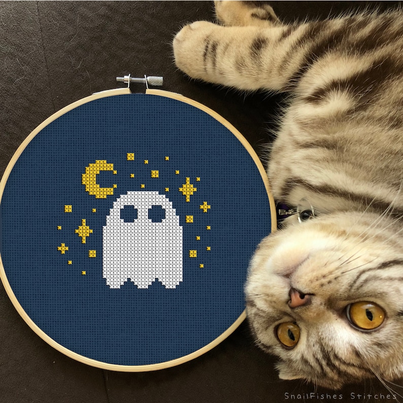 Op de afbeelding: Een blauwe borduurring met een witte geest en gouden sterren en een halve maan op een donkerblauwe achtergrond. De ring ligt op een bruin oppervlak met een tabby-kat die ernaar kijkt.