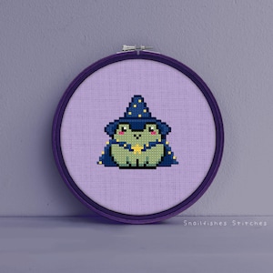 May include: A purple embroidery hoop with a cross-stitch design of a green frog wearing a blue wizard hat with yellow stars. The frog is holding a yellow star in its hand. The background is a light purple fabric.