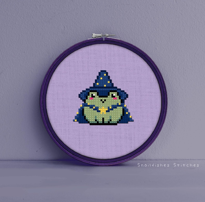May include: A purple embroidery hoop with a cross-stitch design of a green frog wearing a blue wizard hat with yellow stars. The frog is holding a yellow star in its hand. The background is a light purple fabric.