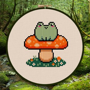 May include: Cross-stitch embroidery of a green frog sitting on a red and orange mushroom. The mushroom is surrounded by green grass and small white flowers. The embroidery is framed in a wooden hoop.